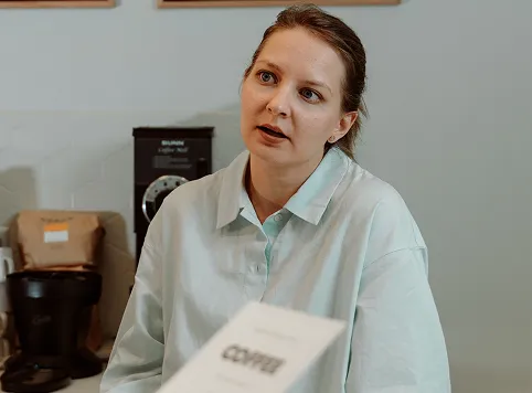 A woman with light-colored hair tied back wearing a pale blue button-up shirt is looking slightly to the side with a thoughtful expression in a room with coffee-making equipment in the background.