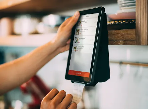 Hands interacting with a touchscreen device mounted on a wall, showing a food ordering app with menu items and an 'Order Out' button, while printing a receipt.