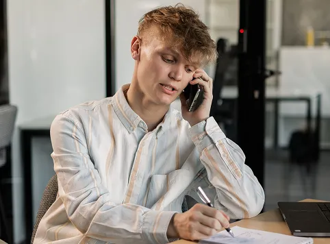 Young man with curly hair sitting at a desk, talking on a smartphone while writing on paper with a pen, with a laptop nearby.