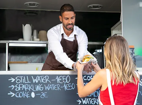 Man in a white shirt and brown apron handing a wrapped sandwich to a woman with blonde hair wearing a red sleeveless top at a food stand with a chalkboard menu listing drinks and prices.