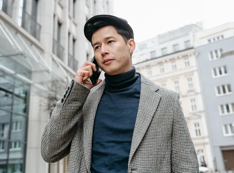 Man wearing a patterned coat and dark turtleneck talking on a smartphone outdoors in an urban setting.