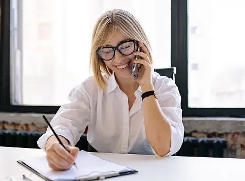 Smiling young woman wearing glasses and a white shirt talking on a smartphone while writing in a notebook at a desk near a window.
