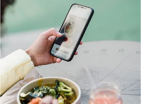A person holding a smartphone and taking a photo of a bowl of food on a gray stone table outdoors, with a glass of pink beverage nearby.