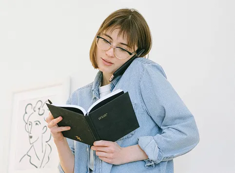 Woman wearing glasses and a denim shirt, holding a notebook and a pen while talking on a phone held between her shoulder and ear.