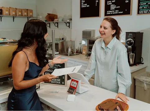 Customer with long dark hair in a dark blue satin dress holding a menu and speaking to a smiling female barista with brown hair tied back, standing behind a white counter in a coffee shop with menu boards and coffee equipment in the background.