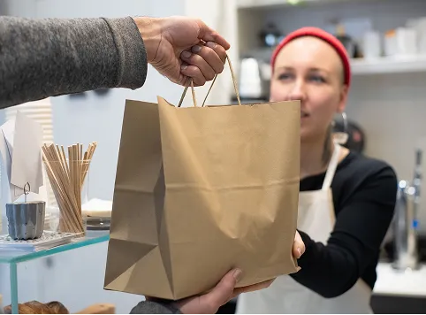 A person handing over a brown paper bag to a woman wearing a red headband and white apron in a cafe or store setting.