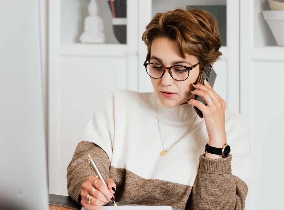 Woman with short hair and glasses talks on phone while writing at a desk with white shelves behind her.