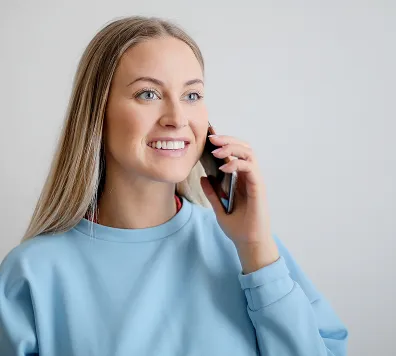Smiling young woman with long blonde hair wearing a light blue sweatshirt, talking on a smartphone against a plain light background.