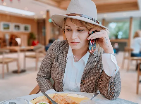 A woman wearing a hat and a checked blazer sits at a table in a restaurant, holding a phone to her ear and looking down at her plate of food with a thoughtful expression.