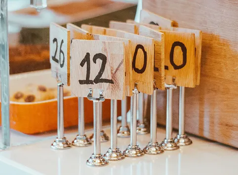 Wooden table number signs with black numerals held by metal stands, arranged in a row on a white surface near a wooden wall.