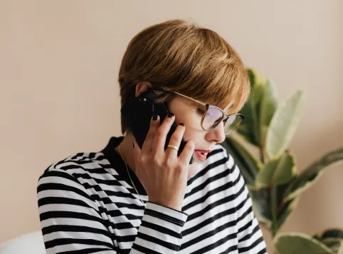 Person with short light brown hair, wearing glasses and a black-and-white striped shirt, talking on a smartphone with a plant in the background.