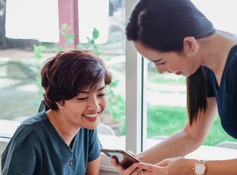 Two women smiling and looking at a smartphone together indoors near a window with greenery outside.
