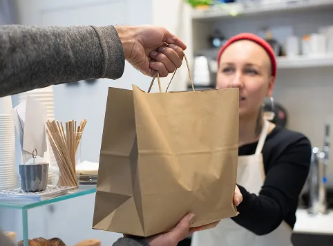 A person hands a brown paper bag to a woman wearing a red beanie and white apron in a cafe or shop setting.