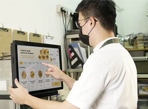 Man wearing a white shirt and black face mask using a touchscreen kiosk to browse and select food delivery options in a kitchen or restaurant setting.