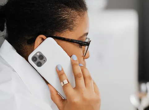 Close-up of a person with glasses holding a white smartphone to their ear while talking, with light blue painted nails and a gold ring on the finger.