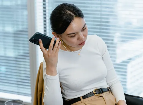 Woman wearing a white long-sleeve top and beige pants sitting indoors near a window, holding a smartphone up to her ear as if talking or listening.