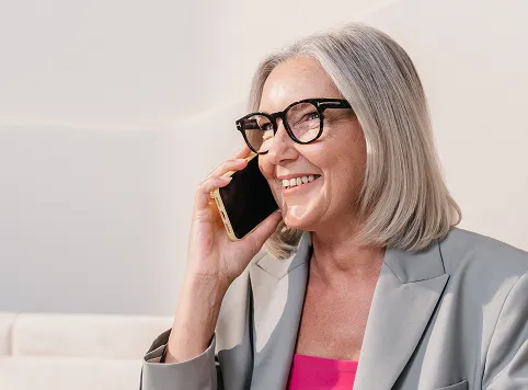 Smiling older woman with gray hair and black-framed glasses talking on a smartphone, wearing a gray blazer and pink top.
