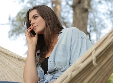 Young woman in a light blue shirt sitting in a hammock outdoors, talking on a cellphone with a thoughtful expression.