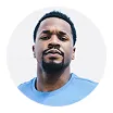 Portrait of a serious young Black man with short hair and a mustache, wearing a light blue shirt against a white circular background.