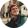 Smiling young man wearing glasses and a beanie outdoors in an urban setting.