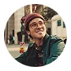 Smiling man wearing glasses, a red beanie, and a green jacket sitting outside in an urban setting.