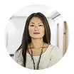 Portrait of a young woman with long dark hair wearing a light-colored sweater and a necklace, standing in a bright hallway.