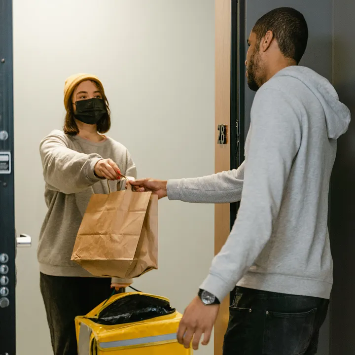 Person wearing a black face mask and yellow beanie handing over a brown paper bag to another person outside a doorway, with a yellow delivery bag on the floor nearby.