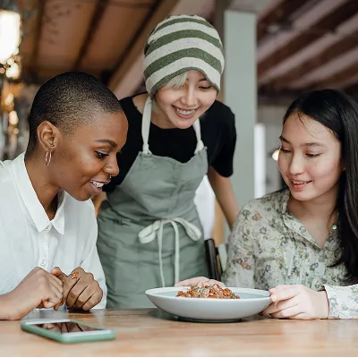Three women smiling and looking at a plate of food on a wooden table; one woman wears a striped headscarf and apron, indicating a food service or café setting.