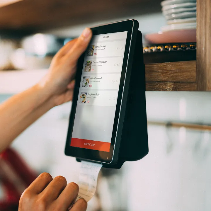 Hand holding a receipt from a touchscreen point-of-sale device mounted on a wall displaying a food order including Curried Swiss, Chicken Chop Suey, Crispy Seaweed, and King Prawn Balls with prices and a red 'CHECK OUT' button at the bottom.
