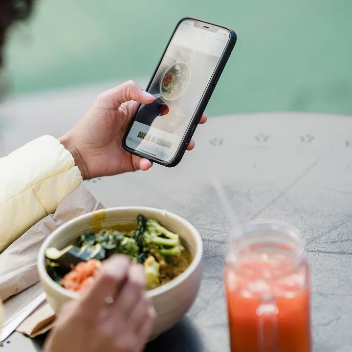 Person holding a smartphone in one hand while eating a bowl of mixed vegetables with the other hand, with a glass of orange juice on the table.