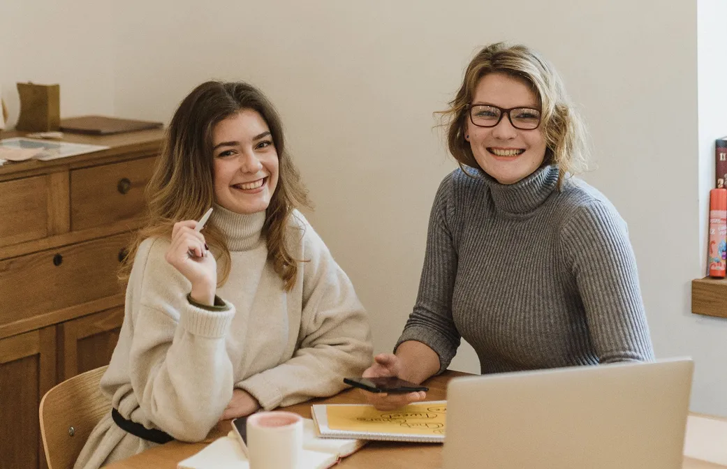 Two smiling women seated at a table with a laptop, phone, and notebooks in a cozy room.