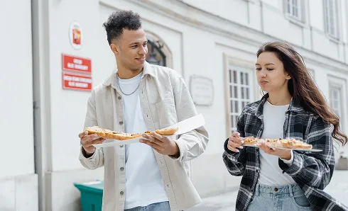 A young man and woman walking outdoors eating slices of pizza from white takeout boxes, with a city building in the background.