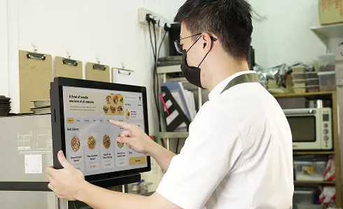 Man wearing glasses and a black face mask, using a touchscreen menu in a kitchen or food preparation area.
