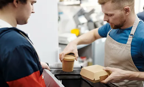 A man in a blue shirt and beige apron hands a cardboard food container and a round container with a lid to another person wearing a dark jacket with red sleeves.