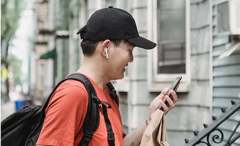 Young man wearing a black cap and red shirt, carrying a black backpack and holding a smartphone while smiling on a city street.