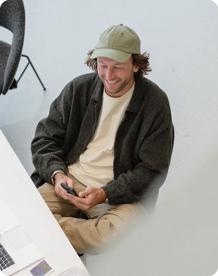 Young man in a green cap and dark sweater smiling while looking at a smartphone in his hands.