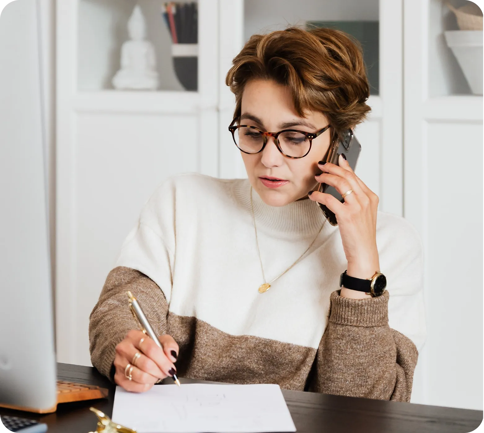 Woman wearing glasses writing on paper while talking on phone at desk with laptop nearby.