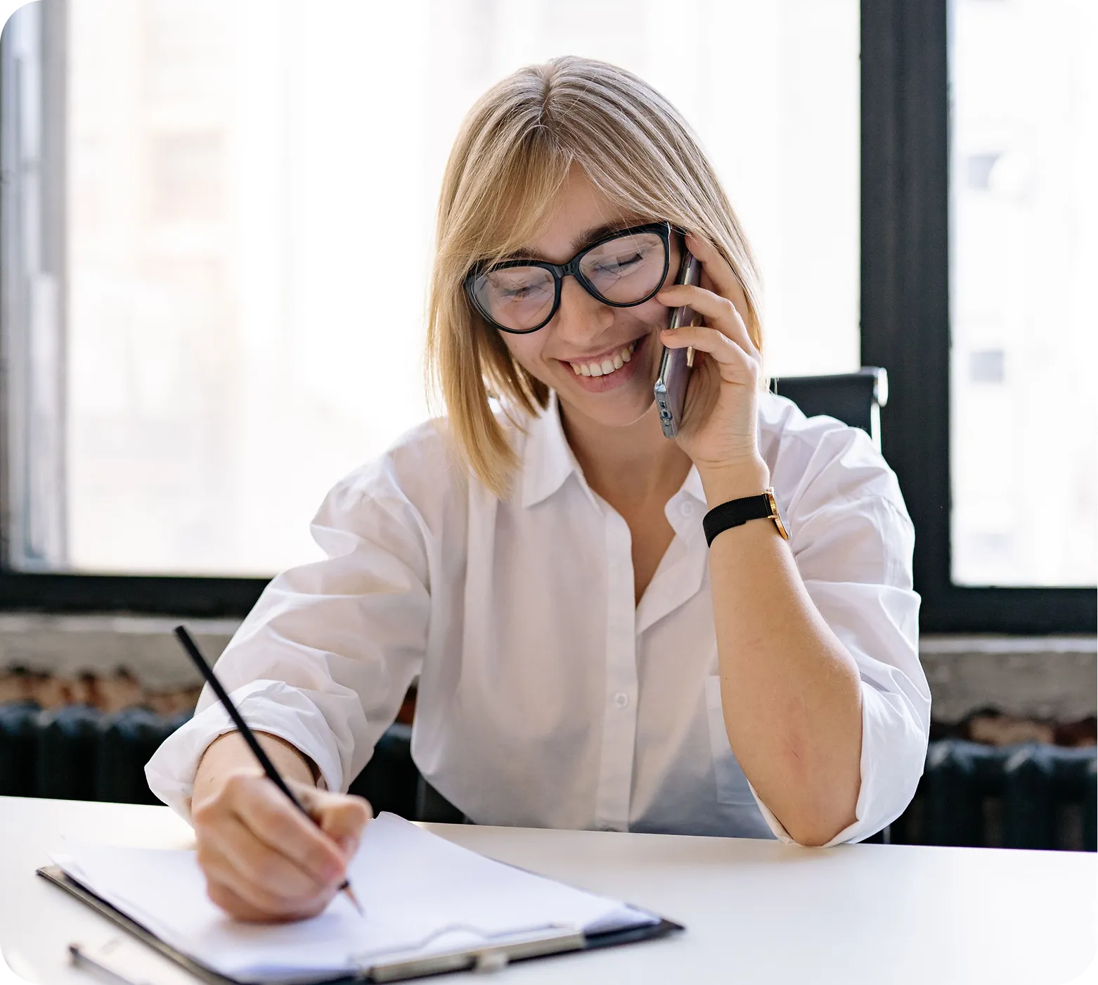 Smiling woman with glasses talks on phone while writing on clipboard in a bright office.