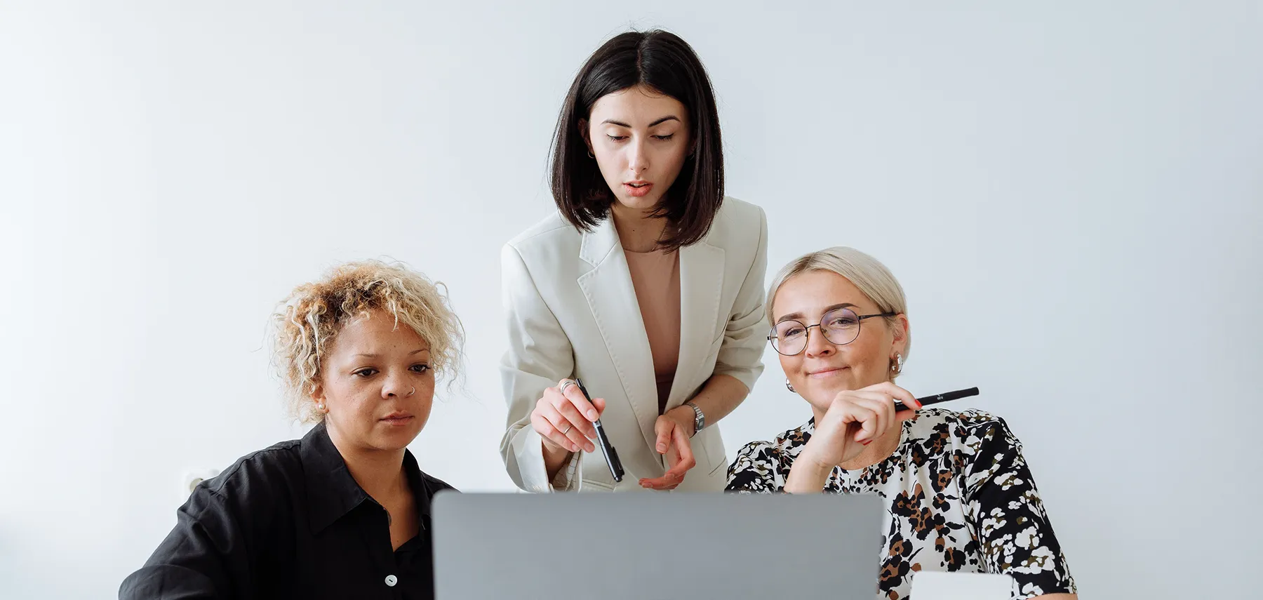 Three women collaborating and looking at a laptop screen in a bright office space.