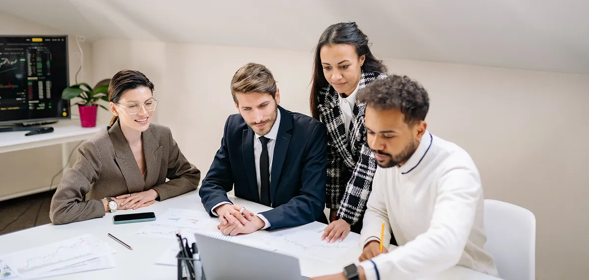 Four diverse business professionals in a meeting reviewing charts and a laptop at a white table.
