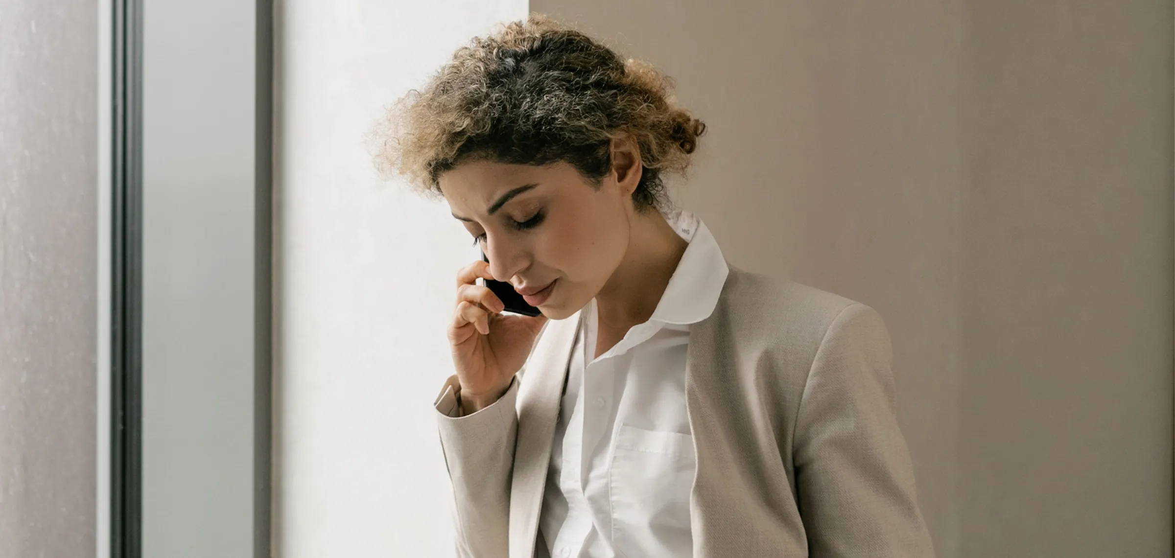 Woman with curly hair in a beige blazer and white shirt talking on a mobile phone near a window.