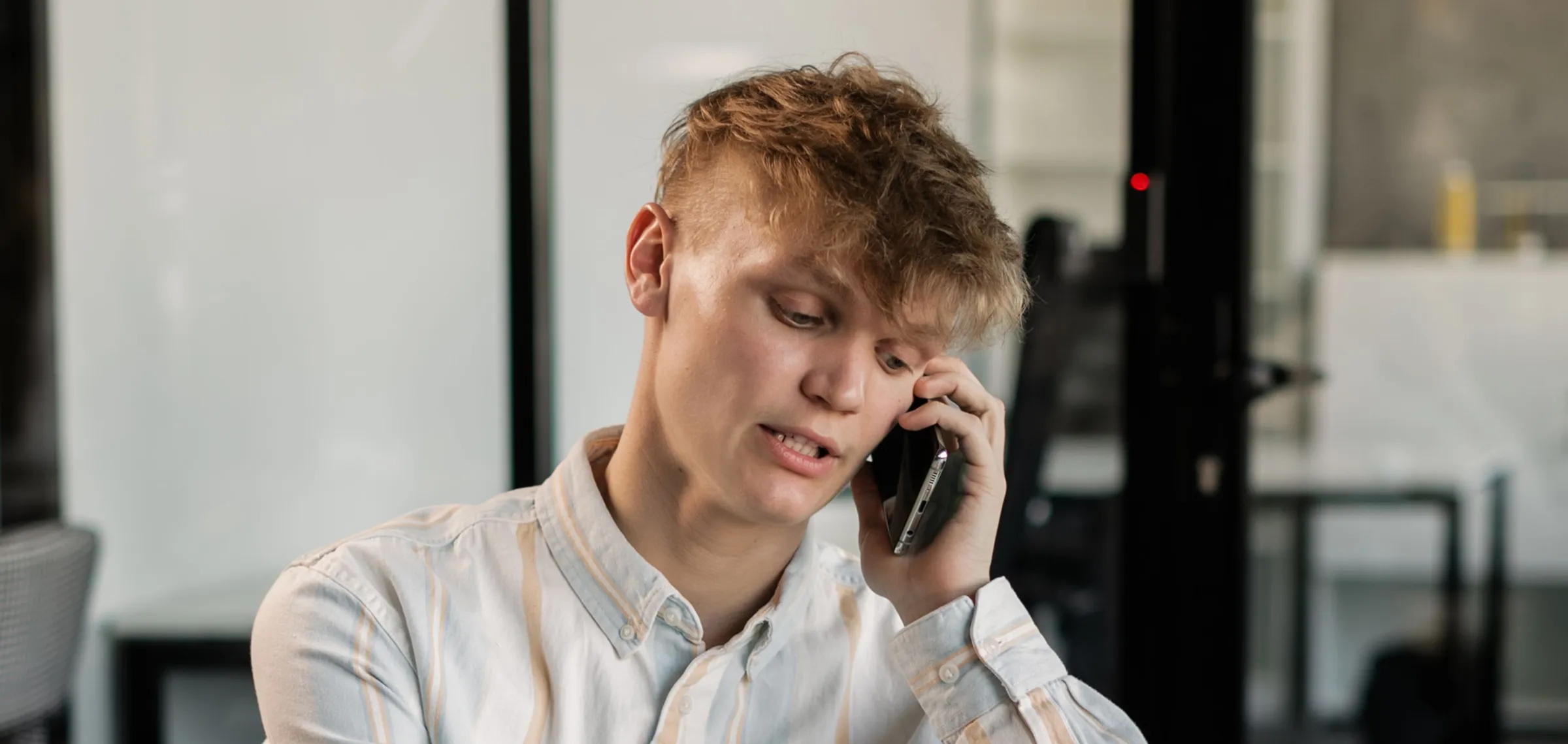 Young man with light brown hair wearing a light-colored button-up shirt talking on a smartphone indoors, with a blurred modern office background.