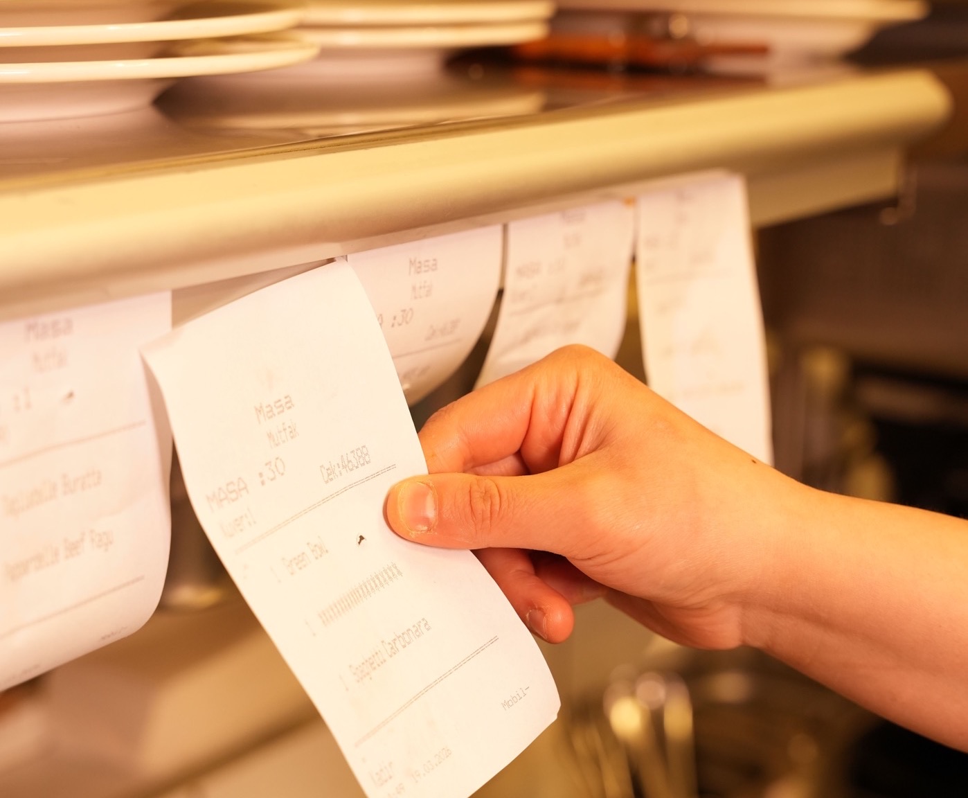 Une main attrape un ticket sur la rampe d'une cuisine pleine de commandes