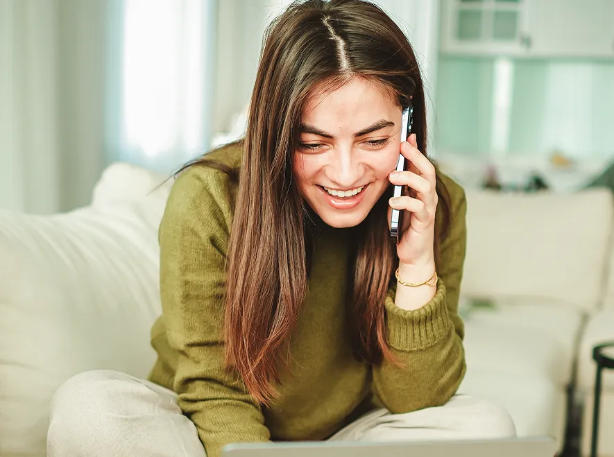 Smiling young woman with long brown hair wearing a green sweater talking on a smartphone while looking at a laptop, seated on a couch in a bright room.