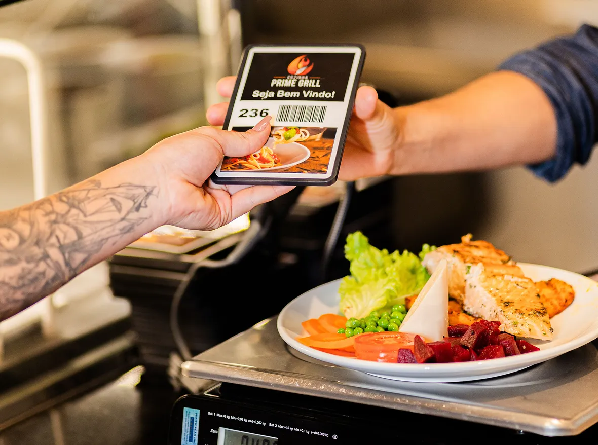 A tattooed hand holding a small card with the text PRIME GRILL and a barcode, handing it over a counter where a plate with grilled chicken breast, lettuce, peas, sliced carrots, tomatoes, and beets is placed on a digital scale.