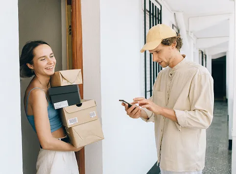 A woman standing in a doorway holding several packages and smiling, while a man wearing a beige cap and shirt uses his phone nearby in a hallway with white walls and tiled floor.