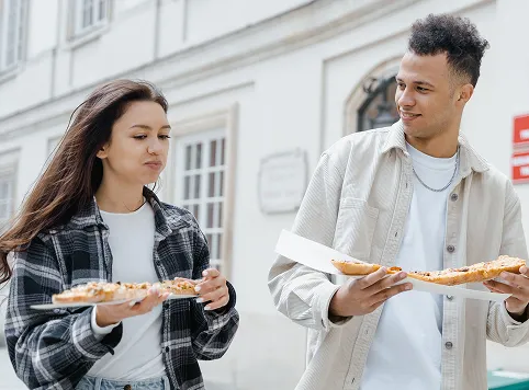 A young woman and a young man outdoors holding and eating slices of pizza while walking near a building with white walls and windows.