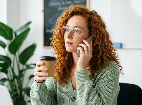 Young woman with curly red hair and glasses holding a coffee cup in one hand and a phone to her ear, sitting indoors with a plant and a blurred chalkboard in the background.