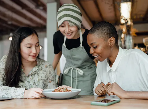 Three women gathered around a table, smiling and looking at a plate of food, with one wearing a green and white striped beanie and apron.